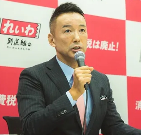 れいわ新選組の山本太郎代表が記者会見でマイクを手に発言する様子、「消費税は廃止!」の背景パネルあり