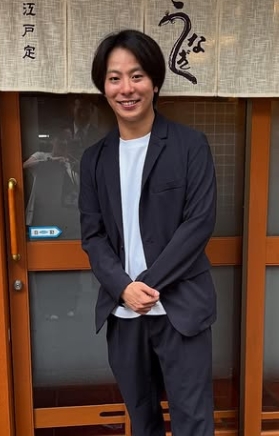中村鶴松さんがカジュアルなスーツ格好ででうなぎ屋の店先で立っている姿